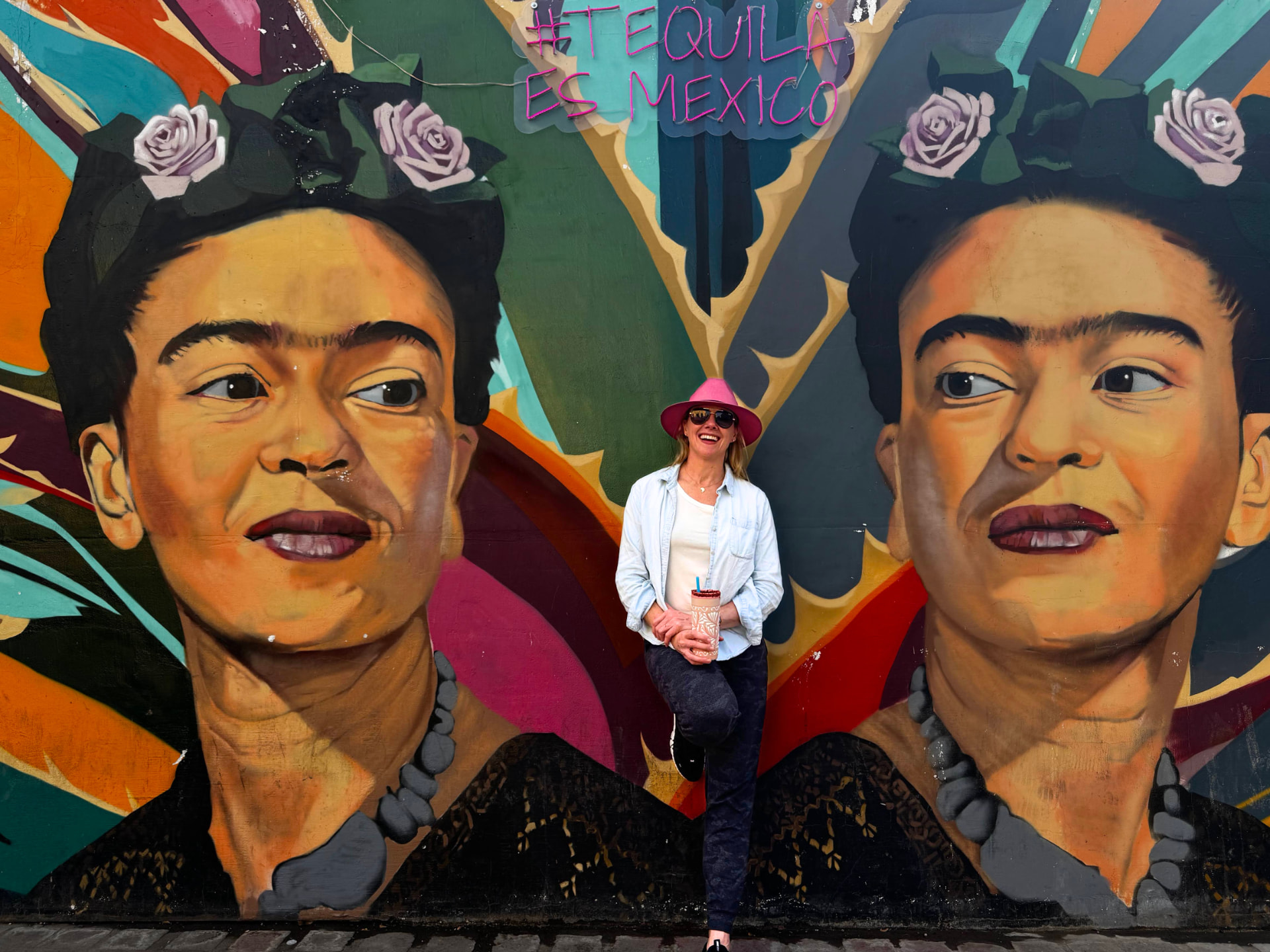 Susan Sjolund in a pink hat smiling in front of a colorful Frida Kahlo mural
